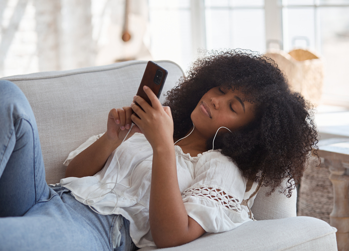 A calm young black lady using wireless headphones, reclining on a comfy sofa, listening to music, audiobooks, or podcasts, and enjoying stress-free holiday recreational time at home.